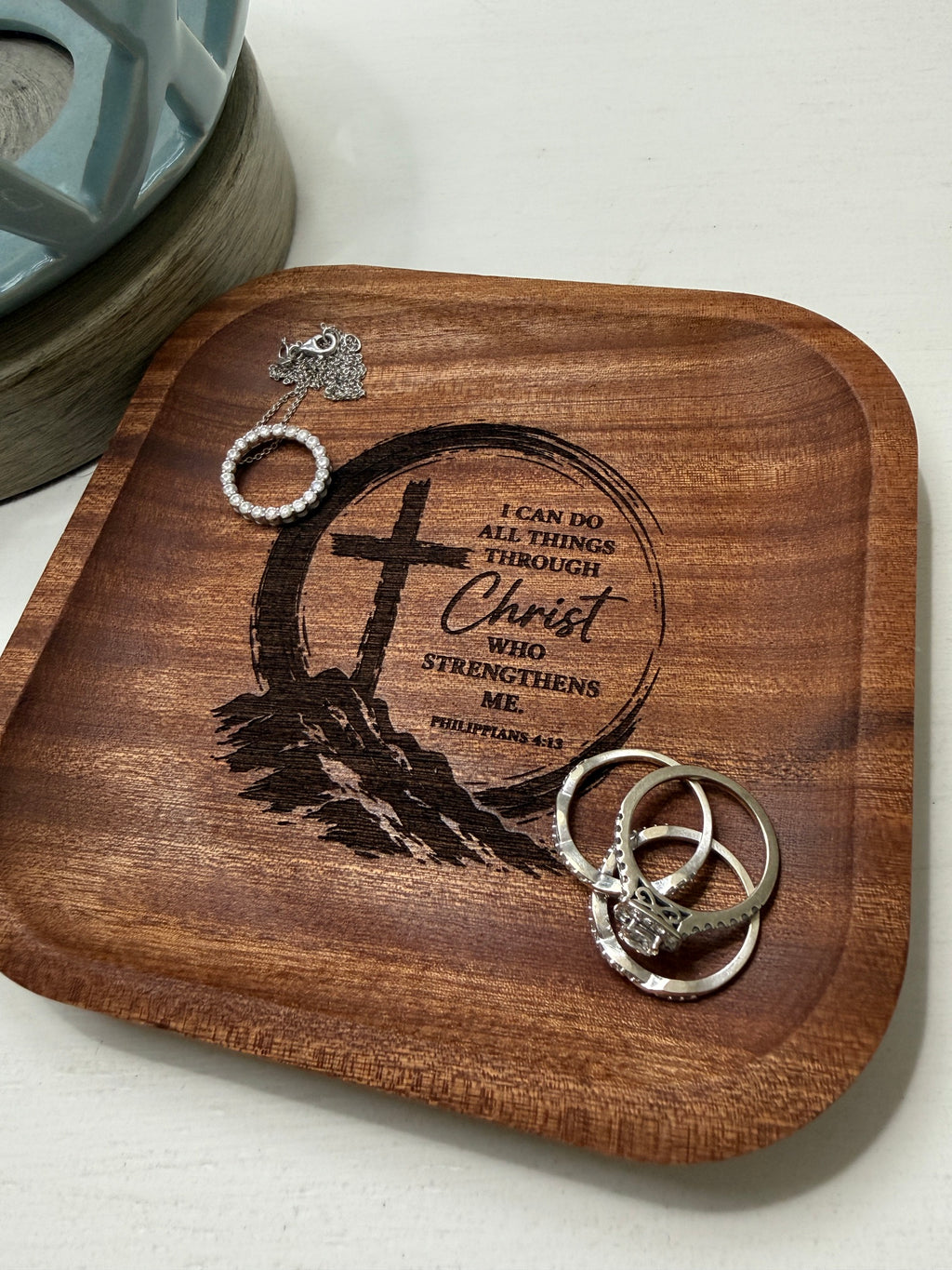 A square wooden tray with rounded corners, engraved with a Christian cross, feather, and the words 'Christ Strengthen Me,' holding several silver rings and a small pearl-and-silver charm against a light wall.
