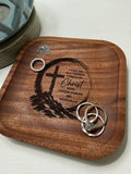 A square wooden tray with rounded corners, engraved with a Christian cross, feather, and the words 'Christ Strengthen Me,' holding several silver rings and a small pearl-and-silver charm against a light wall.